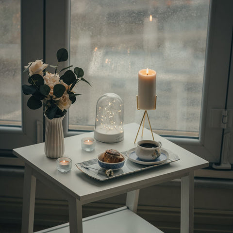 A cozy indoor scene by a window with a small white side table holding a vase of flowers, a lit candle on a metal stand, a glass dome lamp, a cup of coffee, and a pastry. Part of a dark chair with a white throw blanket is visible in the foreground.